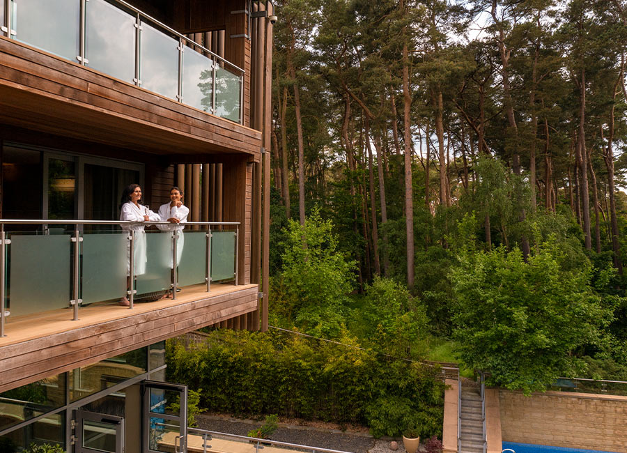 Two women standing on their Spa Suite balcony looking out over the Outdoor Pool to the surrounding forest.